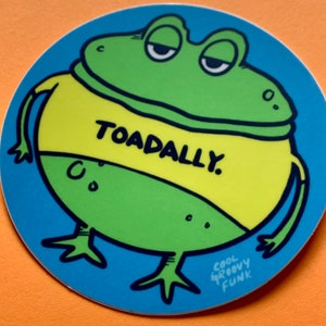 May include: A green frog wearing a yellow shirt with the text "TOADALLY." printed on it. The frog is standing on a blue background with the text "COOL GROOVY FUNK" in white.