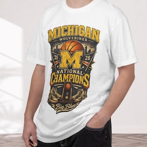 Puede incluir: Camiseta blanca con un gráfico desgastado. El diseño incluye el texto "MICHIGAN WOLVERINES NATIONAL CHAMPIONS", una pelota de baloncesto, la letra "M" y los años "20" y "26". También están presentes las palabras "Big Blue".
