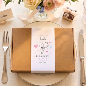 May include: A rectangular brown cardboard activity box with a white paper sleeve featuring the text "OUR SPECIAL GUEST Amelia ACTIVITY BOX THE WEDDING OF STEVE & KELLY 11.07.2024" and a cartoon illustration of a couple holding hands with pink and blue hearts.