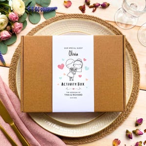 May include: A rectangular brown cardboard box with a white paper band featuring the text "OUR SPECIAL GUEST Olivia ACTIVITY BOX THE WEDDING OF TINA & RICHARD 23.07.2024" and a line drawing of a couple surrounded by small pink and blue hearts.