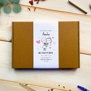 May include: A rectangular brown cardboard box with a white paper band featuring the text "OUR SPECIAL GUEST Amelia ACTIVITY BOX THE WEDDING OF STEVE & KELLY 12.05.2023" and a line drawing of a couple surrounded by small pink and blue hearts.