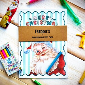 May include: A Christmas activity pack with a Santa Claus illustration, the text "Merry Christmas," and "Freddie's Christmas Activity Pack." The image also includes a box of mini colored pencils, a yellow figure, and various colored pencils.