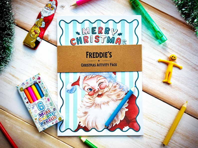 May include: A Christmas activity pack with a Santa Claus illustration, the text "Merry Christmas," and "Freddie's Christmas Activity Pack." The image also includes a box of mini colored pencils, a yellow figure, and various colored pencils.
