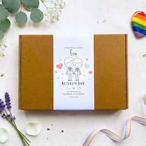 May include: A rectangular brown cardboard box with a white paper band featuring the text Our Special Guest Erin Activity Box The Wedding of George & Patrick 23rd July 2026 and a line drawing of two figures holding hands surrounded by small hearts.
