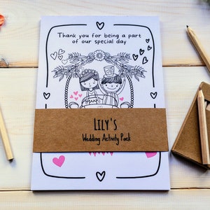 May include: A white colouring book with a brown paper band that says "LILY'S Wedding Activity Pack". The cover features a black and white illustration of a bride and groom in a wedding carriage with hearts and flowers. The book comes with a pack of coloured pencils.