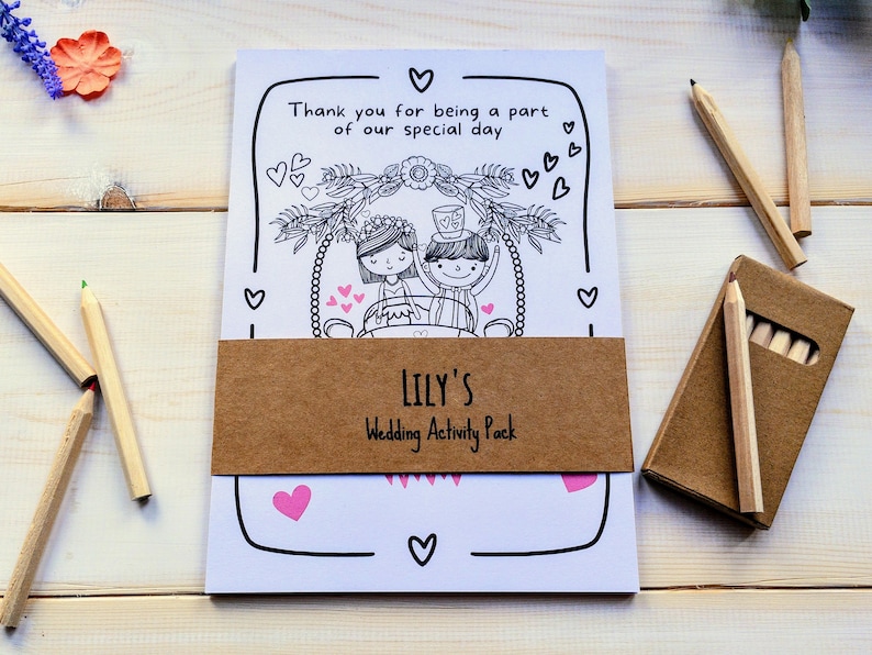 May include: A white colouring book with a brown paper band that says "LILY'S Wedding Activity Pack". The cover features a black and white illustration of a bride and groom in a wedding carriage with hearts and flowers. The book comes with a pack of coloured pencils.