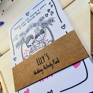May include: A brown paper envelope with a white card inside. The card has a black and white illustration of a bride and groom on a swing. The envelope has the text "LILY'S Wedding Activity Pack" printed on it.