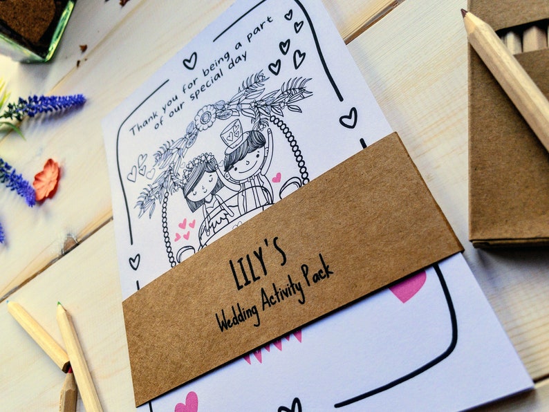 May include: A brown paper envelope with a white card inside. The card has a black and white illustration of a bride and groom on a swing. The envelope has the text "LILY'S Wedding Activity Pack" printed on it.