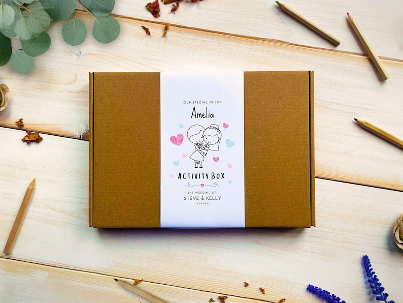 May include: A rectangular brown cardboard box with a white paper band featuring the text "OUR SPECIAL GUEST Amelia ACTIVITY BOX THE WEDDING OF STEVE & KELLY 12.05.2023" and a line drawing of a couple surrounded by small pink and blue hearts.