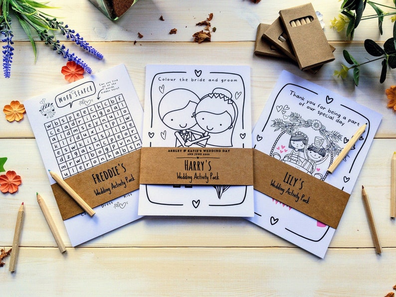 May include: Three wedding activity packs with brown paper wrappers. The packs are labelled "Freddie's", "Harry's", and "Lily's" and include colouring pages and a word search puzzle. The packs are on a light wood surface with coloured pencils.