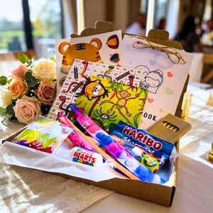 May include: A brown cardboard activity box containing a numbered game board, a colorful monkey-themed puzzle, a white activity book, a multi-colored silicone bubble fidget toy, a pack of Haribo Starmix, Love Hearts candy, and a small bag of dice.