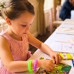 May include: A child wearing a sleeveless pink lace dress with a tulle skirt, adorned with a small floral hair accessory. The child has two neon plastic bracelets on their wrist and is assembling a colorful jigsaw puzzle on a white table.