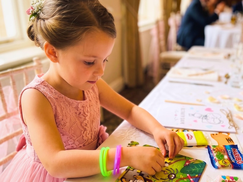 May include: A child wearing a sleeveless pink lace dress with a tulle skirt, adorned with a small floral hair accessory. The child has two neon plastic bracelets on their wrist and is assembling a colorful jigsaw puzzle on a white table.