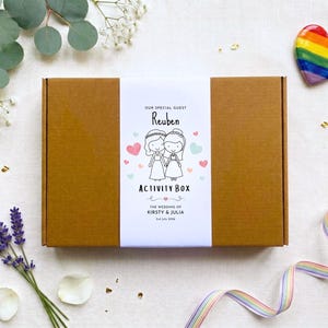 May include: A rectangular brown cardboard box with a white paper band featuring a cartoon illustration of two people in wedding attire, the text "OUR SPECIAL GUEST REUBEN ACTIVITY BOX" and "THE WEDDING OF KIRSTY & JULIA 2nd July 2024" in black ink.