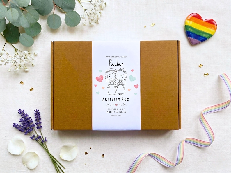 May include: A rectangular brown cardboard box with a white paper band featuring a cartoon illustration of two people in wedding attire, the text "OUR SPECIAL GUEST REUBEN ACTIVITY BOX" and "THE WEDDING OF KIRSTY & JULIA 2nd July 2024" in black ink.