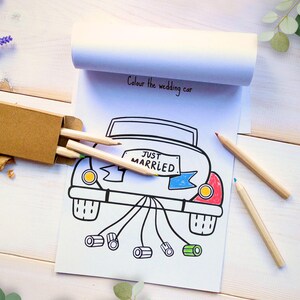 May include: A white paper coloring pad featuring a black line drawing of a car with the text "JUST MARRIED" on the license plate and "Colour the wedding car" at the top. A small brown cardboard box contains wooden pencils next to the pad.