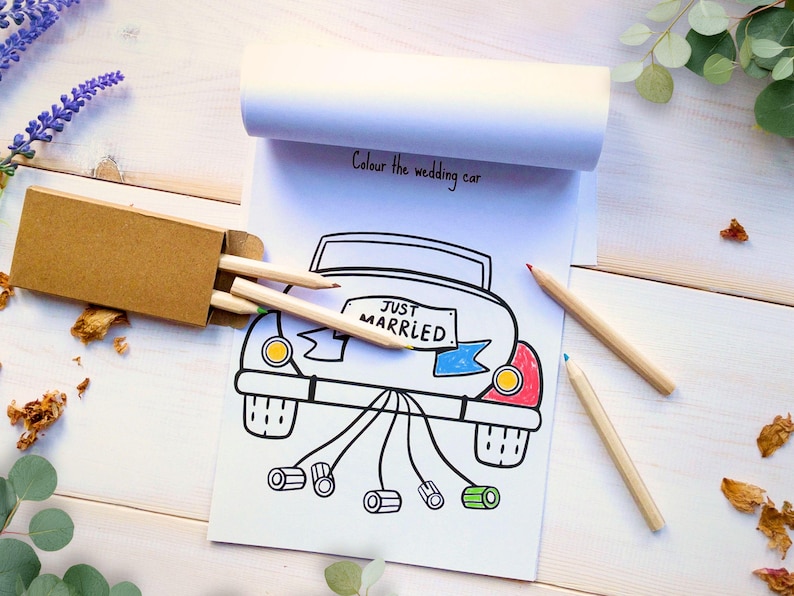 May include: A white paper coloring pad featuring a black line drawing of a car with the text "JUST MARRIED" on the license plate and "Colour the wedding car" at the top. A small brown cardboard box contains wooden pencils next to the pad.