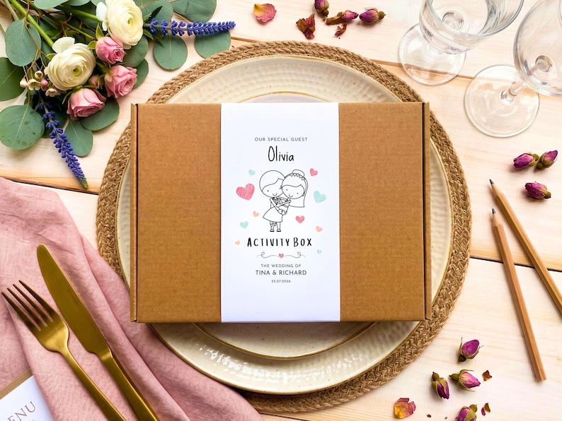 May include: A rectangular brown cardboard box with a white paper band featuring the text "OUR SPECIAL GUEST Olivia ACTIVITY BOX THE WEDDING OF TINA & RICHARD 23.07.2024" and a line drawing of a couple surrounded by small pink and blue hearts.