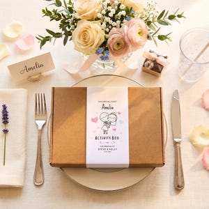 May include: A rectangular brown cardboard activity box with a white paper sleeve featuring the text "OUR SPECIAL GUEST Amelia ACTIVITY BOX THE WEDDING OF STEVE & KELLY 11.07.2024" and a cartoon illustration of a couple holding hands with pink and blue hearts.