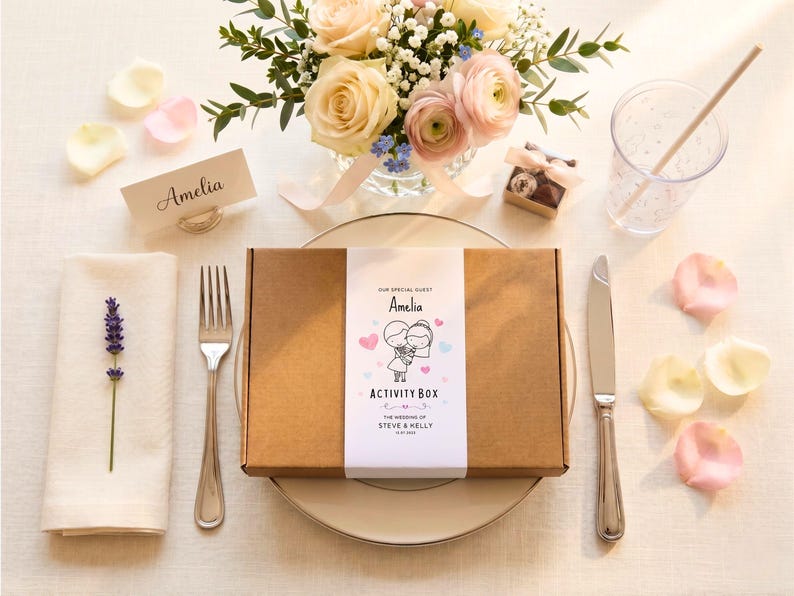 May include: A rectangular brown cardboard activity box with a white paper sleeve featuring the text "OUR SPECIAL GUEST Amelia ACTIVITY BOX THE WEDDING OF STEVE & KELLY 11.07.2024" and a cartoon illustration of a couple holding hands with pink and blue hearts.