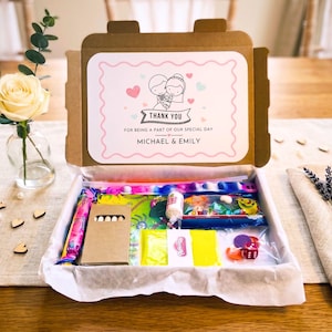 May include: A cardboard gift box containing assorted small toys and treats. The inside lid features a printed label with a cartoon illustration of a couple, pink hearts, and the text: THANK YOU FOR BEING A PART OF OUR SPECIAL DAY MICHAEL & EMILY.