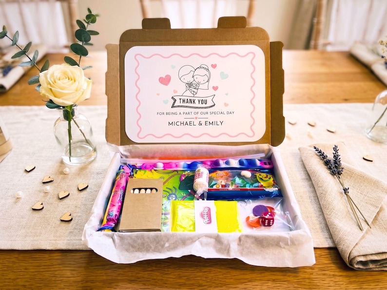 May include: A cardboard gift box containing assorted small toys and treats. The inside lid features a printed label with a cartoon illustration of a couple, pink hearts, and the text: THANK YOU FOR BEING A PART OF OUR SPECIAL DAY MICHAEL & EMILY.