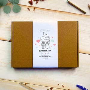 May include: A rectangular brown cardboard box featuring a white paper belly band. The band displays the text "OUR SPECIAL GUEST Erin ACTIVITY BOX" and "THE WEDDING OF GEORGIA & PATRICK 23rd May 2026" with a cartoon illustration of a bride and groom.