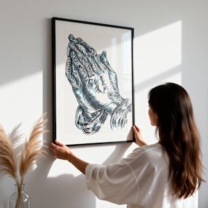 K&ouml;nnte beinhalten: Ein gerahmter Wandkunstdruck mit einer detaillierten Illustration von zwei in Gebetshaltung zusammengelegten H&auml;nden. Das Kunstwerk verwendet kr&auml;ftige schwarze Umrisse und blaue Schattierungen auf wei&szlig;em Grund in einem schwarzen Rahmen.