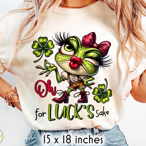 Op de afbeelding: Crèmekleurig T-shirt met een cartoonkikker met een rode strik, knipogend en met een bruine korte broek en laarzen. De tekst "Oh for LUCK'S sake" staat onder de kikker, met klavers aan weerszijden. Het shirt meet 38 x 46 cm.