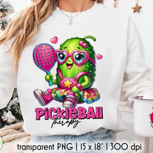 Op de afbeelding: Witte sweatshirt met een cartoon augurkenpersonage met een roze bril en een pickleball-racket. Het ontwerp bevat de tekst "Pickleball Therapy" in het roze. De afbeelding bevat ook een transparante PNG van 38 x 46 cm.