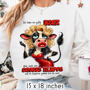 Może przedstawiać: Biała bluza z kapturem z kreskówkową krową z blond kręconymi włosami, czerwonymi ustami i czerwoną sukienką. Niemiecki tekst brzmi: "Mam dobre serce, ale także duży pysk i jestem trochę złośliwa". Mierzy 38 x 46 cm.