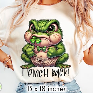 Puede incluir: Camiseta color crema con una rana de dibujos animados con expresión enfadada, sosteniendo un trébol de cuatro hojas. La rana lleva pajarita y pañal. El texto en la camiseta dice "I PINCH BACK!" La camiseta mide 38 x 46 cm.