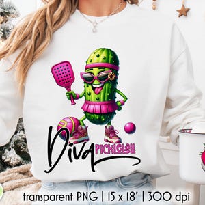 Op de afbeelding: Witte sweatshirt met een cartoon augurkenpersonage met een zonnebril, een roze vizier en een rok, met een paddle en staand bij een bal. De tekst "Diva Pickleball" is geschreven in zwart script. Transparante PNG.