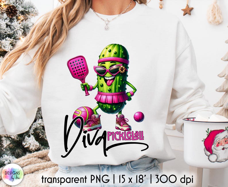 Op de afbeelding: Witte sweatshirt met een cartoon augurkenpersonage met een zonnebril, een roze vizier en een rok, met een paddle en staand bij een bal. De tekst "Diva Pickleball" is geschreven in zwart script. Transparante PNG.