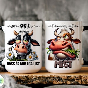 May include: Two white ceramic mugs with black handles and rims. Each mug features a cartoon cow illustration with text. One cow has daisies and the text reads "DASS ES MIR EGAL IST". The other cow has the text "MIST".