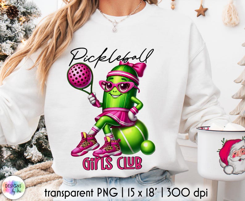Op de afbeelding: Witte sweatshirt met een cartoon augurkenpersonage dat pickleball speelt. De augurk draagt een roze bril, een hoofdband en een rok, met de woorden "Pickleball Girls Club" in het roze. Het ontwerp bevat een pickleball-racket en -bal.