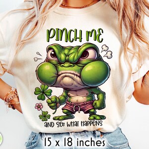 May include: Cream-coloured t-shirt with a cartoon frog. The green frog has an angry expression and wears pink shorts, holding a four-leaf clover. Text on the shirt reads "PINCH ME" and "AND SEE WHAT HAPPENS". The shirt measures 38 x 46 cm.