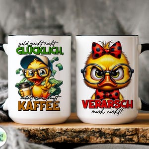 May include: Two white ceramic mugs with black handles and rims. One mug features a cartoon duck with glasses, a hat, and the text "geld macht nicht GL&Uuml;CKLICH, au&szlig;er man kauft KAFFEE." The other mug has a duck with a bow tie and the text "VERARSCH mich nicht!"