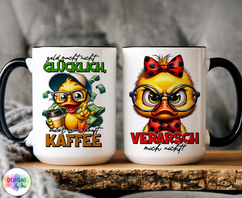May include: Two white ceramic mugs with black handles and rims. One mug features a cartoon duck with glasses, a hat, and the text "geld macht nicht GL&Uuml;CKLICH, au&szlig;er man kauft KAFFEE." The other mug has a duck with a bow tie and the text "VERARSCH mich nicht!"