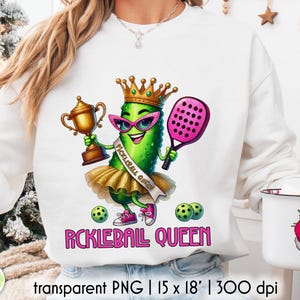 Op de afbeelding: Witte sweatshirt met een cartoon augurkenpersonage met een kroon, zonnebril en een tutu, met een trofee en een roze pickleball-peddel. De tekst "PICKLEBALL QUEEN" wordt in het roze weergegeven. Er worden ook twee pickleballs getoond.