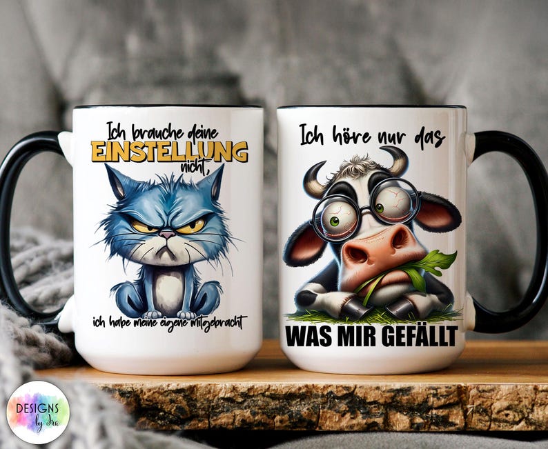 May include: Two white ceramic mugs with black handles and rims. One mug features a grumpy blue cat illustration with German text. The other mug has a cow wearing glasses and eating grass, also with German text. Both mugs have text.