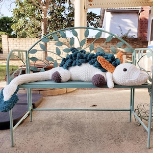 May include: A large, handmade crochet unicorn plushie in white, teal, brown, and orange, resting on a teal metal bench. The unicorn has a long tail, a mane, and a horn. The bench is outdoors.