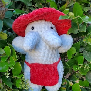 Puede incluir: Un peluche de elefante blanco hecho a mano con ganchillo, con un sombrero rojo en forma de hongo y pantalones cortos rojos. El elefante tiene orejas grandes y esponjosas y una trompa. El juguete está rodeado de hojas verdes.