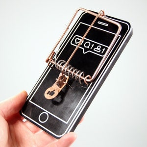 May include: A black smartphone with a copper-colored mouse trap on top. The smartphone screen shows a social media notification with a heart, a comment icon, and the number "1".