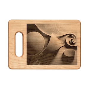 Puede incluir: Tabla de cortar de madera clara con asa. El centro presenta un diseño tallado detallado de formas abstractas, incluyendo una espiral, círculos y un triángulo. La veta de la madera es visible en toda la tabla.