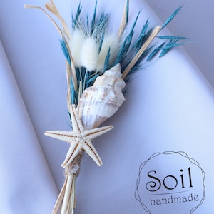 May include: A small bouquet of dried grasses and flowers with a seashell and a starfish. The bouquet is tied with twine and has a white fluffy flower. The text "Soil handmade" is visible in a circle.