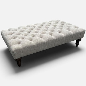 Puede incluir: Un otomano rectangular tapizado en una tela texturizada de color blanco roto con un diseño de capitoné profundo en la superficie superior. La pieza se apoya sobre cuatro patas de madera marrón oscuro con forma torneada y redondeada.