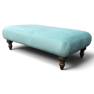 Peut inclure: Un pouf rectangulaire recouvert d'un tissu bleu clair doux à la texture lisse. Ce meuble est doté de quatre pieds en bois brun foncé au design arrondi et tourné, assurant le soutien à chaque coin de la base.