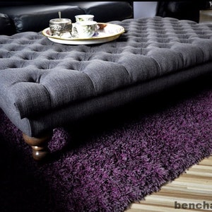 Pode incluir: Um otomano cinzento escuro estofado com pernas de madeira sobre um tapete de pelo roxo.