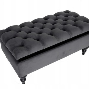 May include: A dark grey upholstered storage ottoman with a tufted top. The rectangular ottoman has a hinged lid and sits on four dark, turned wooden legs. The interior is visible, suggesting storage space.